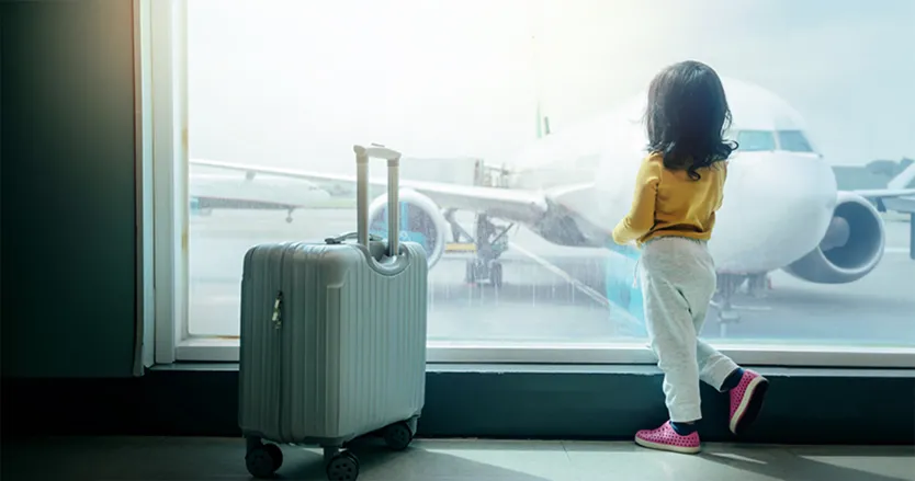 Little girl standing in departure lounge looking out at a plane with a carry on at her side